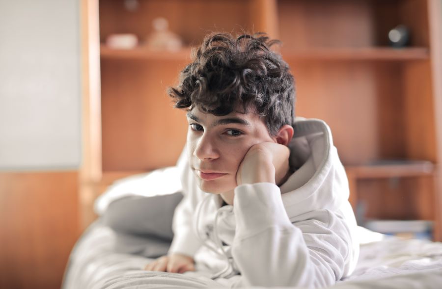 A teenage boy with messy hair wearing a hoodie and leaning his face against his hand while lying in bed.