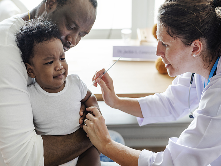 Dad holding grimacing baby in doctor's office, as a nurse gives baby a shot. 