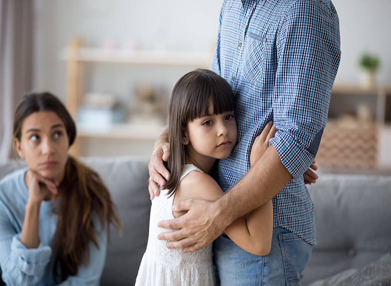 Sad little girl hugs father while mother watches from afar.