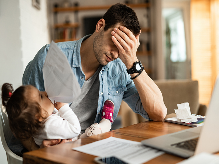 Stressed dad, with his head in his hand, siting at a desk at home trying to work while holding toddler daughter on his lap.